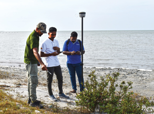 Three men stand by a shore, one points at something on the ground.