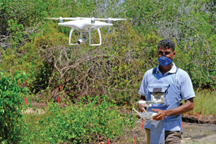A drone hovers in front of a man wearing personal protective equipment and a mask holds a controller for a drone.