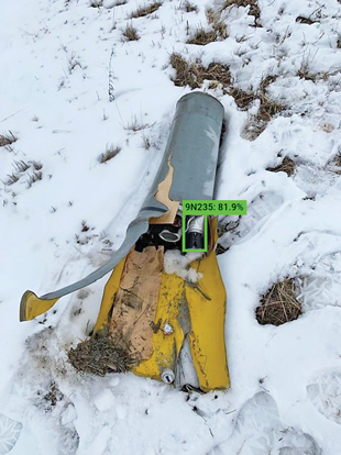 A submunition lying on the snowy ground with a superimposed green box aroind it.