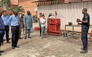 A group of people stand in a dirt courtyard listening to a trainer discuss tools used in demining.