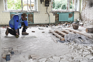 A man kneels and observes something on a concrete floor inside a large room with battle damage.