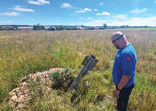A man stands in the grass and looks down at a rocket sticking out of a hole in the field.