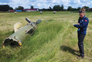 A woman stands in a grassy field next to a large, metal cylinder. Shea writes something on a clipboard.