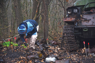 A man in personal protective equipment leans over and looks at something on the ground, in the background is a destroyed tank.