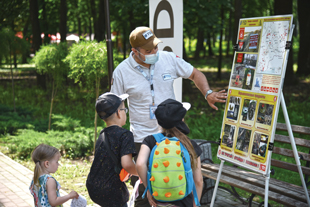 A man in a white shirt and mask talks to children as he points to a display board with pictures of explosive items.