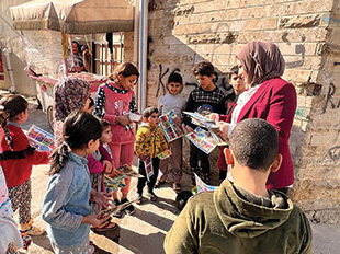 A woman gives handouts to children by a tan-brick wall.