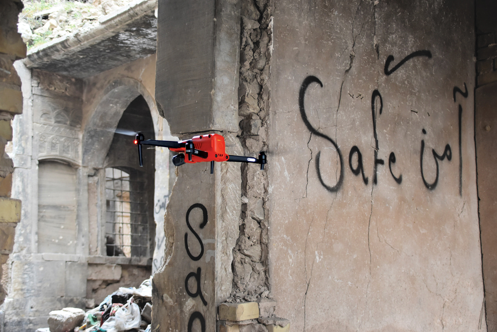 An orange drone flying by ruined buildings with the words "Safe" written in English and Arabic on the wall.