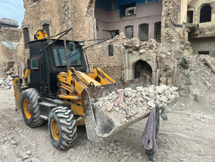 An excavator carrying rubble along a sandy street.