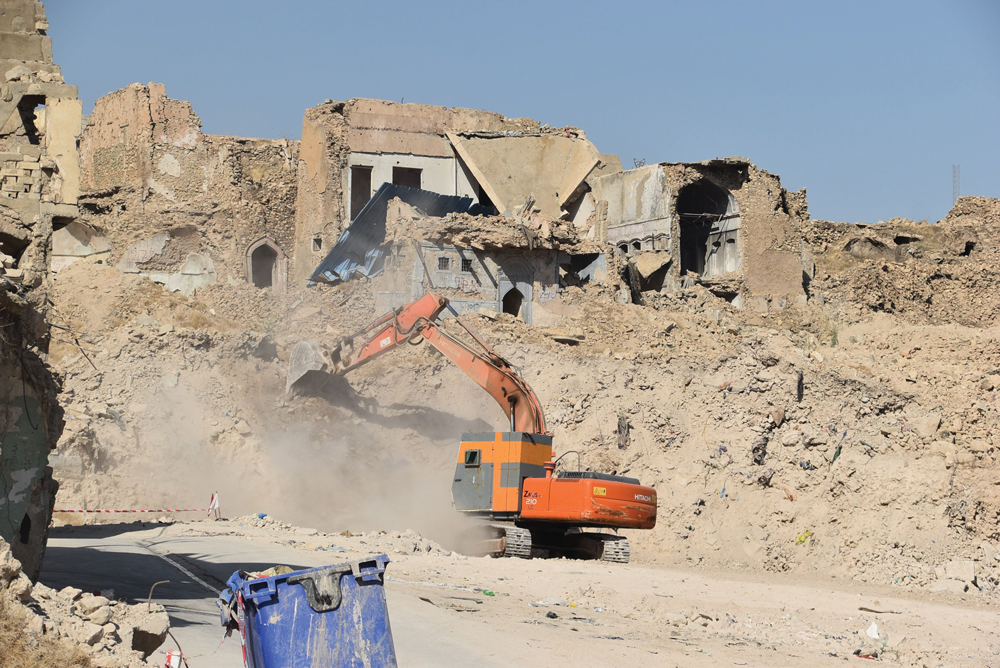 An armored excavator digs rubble from the side of a destroyed building.