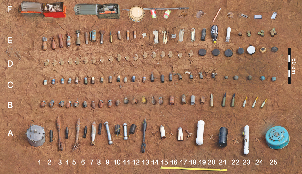 Birdseye view of a dirt field with items of ordnance arranged neatly in four rows.