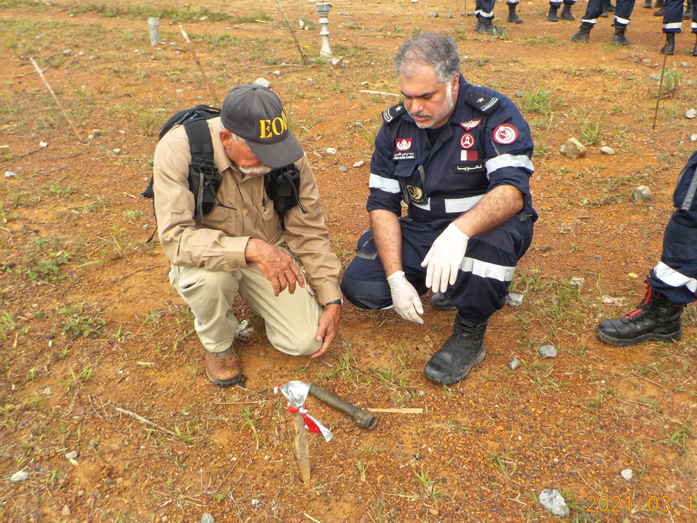 Two men kneel by a small cylinder on the ground.