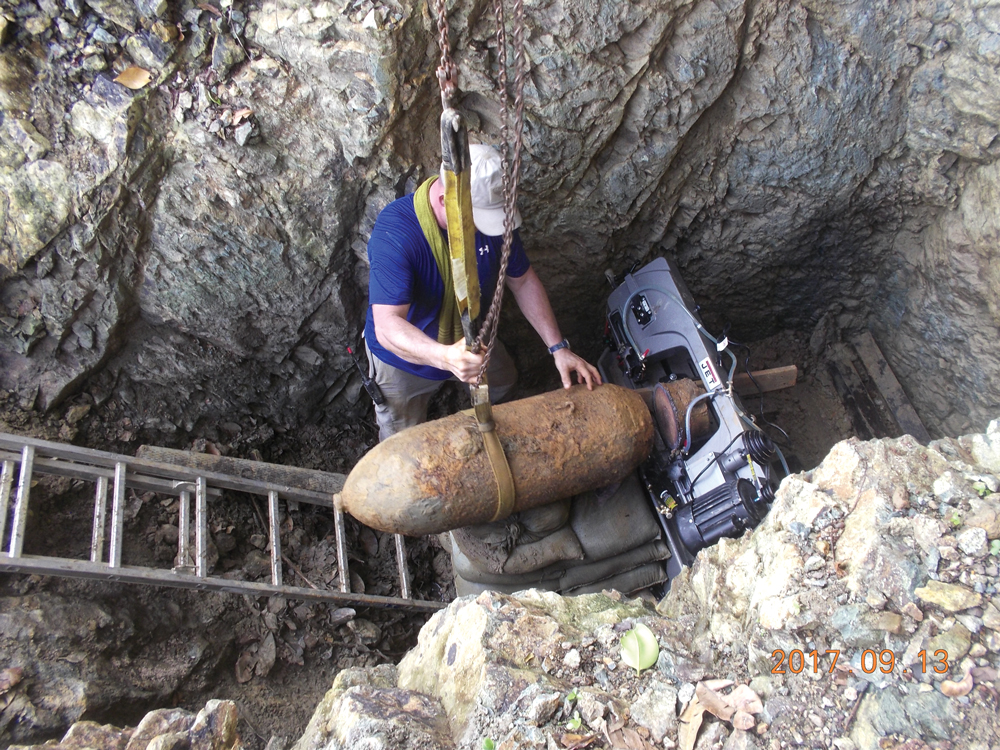 A man standing in a rocky hole with a large rusted piece of ordnance.