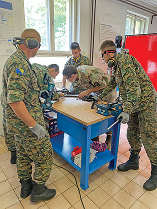 Several men in camouflage uniforms stand around a blue table and work on a firearm component.