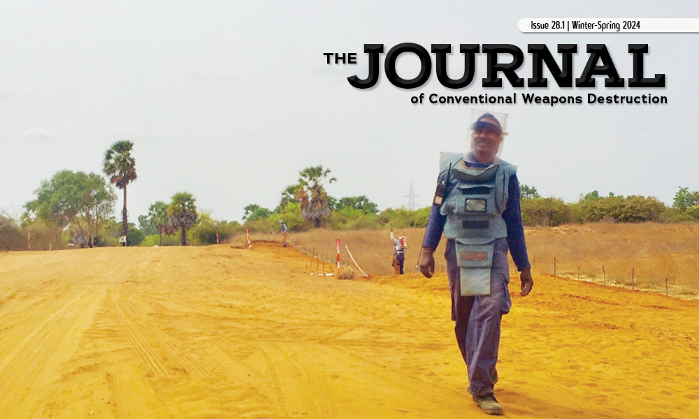 Man in blue personal protective equipment walks across flat, sandy ground while smiling. Super imposed text reads The Journal of Conventional Weapons Destruction.