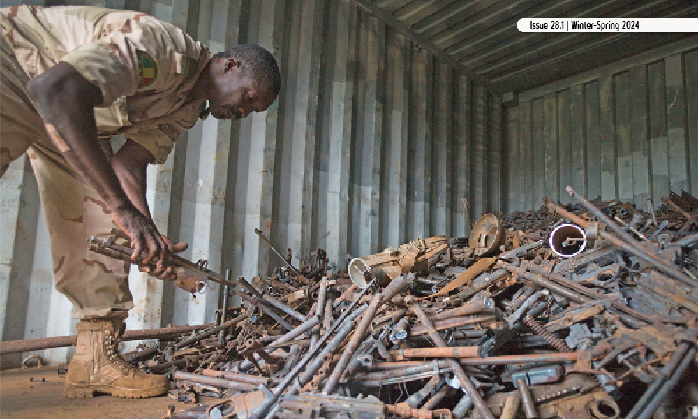 Man in desert fatigues leans over and gathers items from a pile of rusty weapon components.
