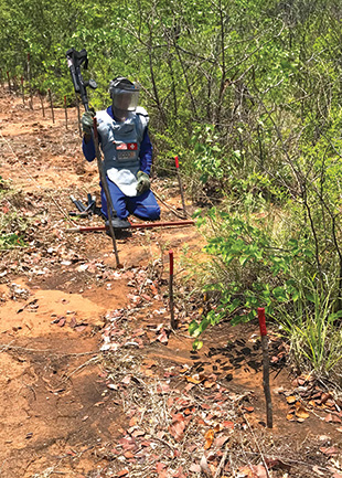 A man in blue protective gear and a metal detector kneels on the ground by short posts.