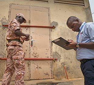 A man in fatigues opens a lock to a metal door and another man in a blue shirt writes on a clipboard.