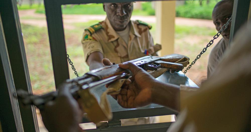 A man hands another man a rifle through metal bars.