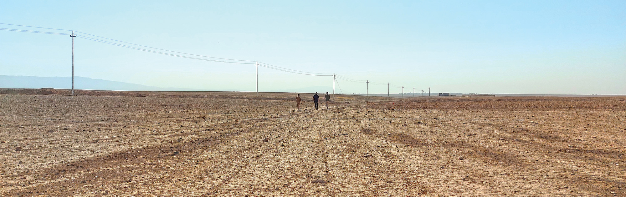 Three people walking side-by-side across desert terrain toward a  