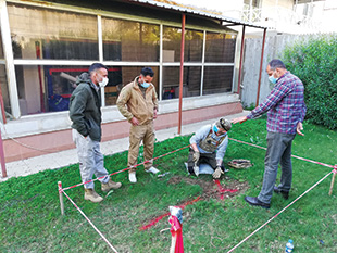 Three men stand by another man kneeling on grass within an enclosure. 