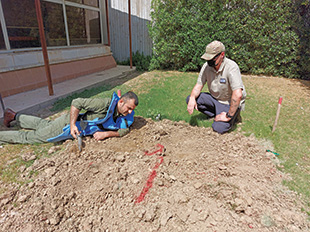 One man wearing a protective vest lies on a dirt pile as another man kneeling nearby observes.
