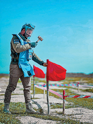 A man in blue personal protective equipment hammers a flag post into the ground.