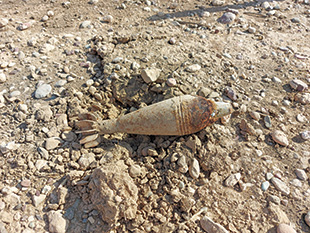 A sand-colored, tear-dropped shaped munition lying on rocky ground.