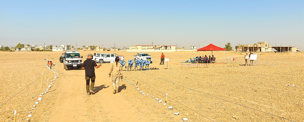 Two people walking along a path marked by posts and tape toward a small camp in the desert.