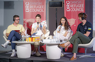 Two women holding microphones and two men wearing headphones, all sitting in white chairs on a stage.