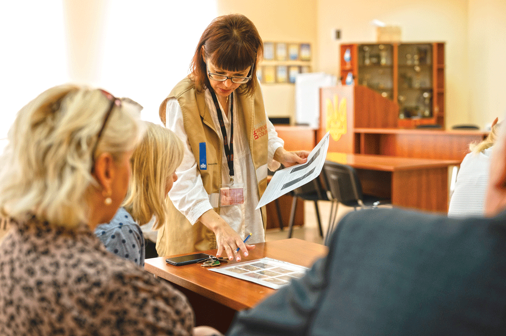 Two women sit and look at a page with pictures, another woman stands and points at the pictures.