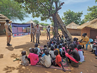 A group of children sit on the ground listening to an adult speak as two other adults hold a large banner that says Local and Improvised Markings.