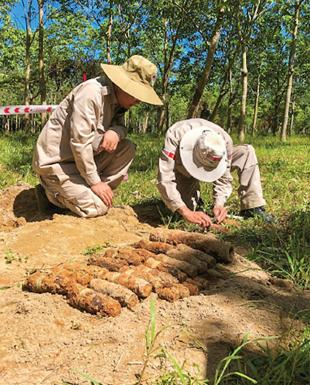 Two people crouch over a collection of rusty metal objects sitting in the dirt.