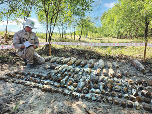 A man wearing beige pants, shirt and hat kneels on the dirt ground next to 6 rows of old dirty rusty metal objects.