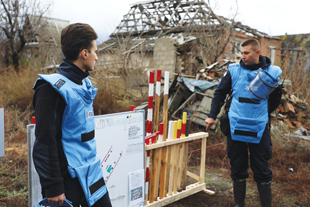 Two people wearing blue protective vests stand outside a damaged building with a rack on wooden stakes with red, white, and yellow painted ends.