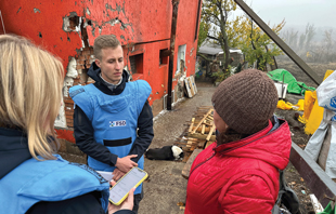 Two people wearing blue protective vests stands in a yard filled with debris talking to a person in a red coat and brown hat.