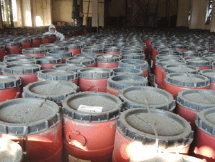A warehouse full of red plastic barrels with black lids. A person wearing a white protective suit stands in the middle.
