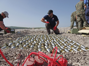 Dozens of metal objects are layed out on the ground close together in a single layer. Two men kneel on the ground on either side of the objects.