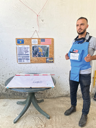 A man wearing a light blue PPE vest that says The HALO Trust stands beside a table with a map on it with a bulletin board hanging on the wall with the flag of Kosovo and the United States, and various maps.