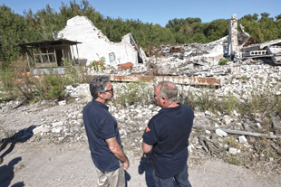 Two men stand talk to each other while facing structures destroyed by bombing.