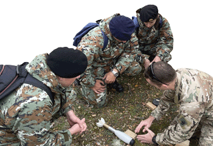 Four men in military fatiques kneel on the ground looking at a 3-D printed projectile used for explosive ordnance training
