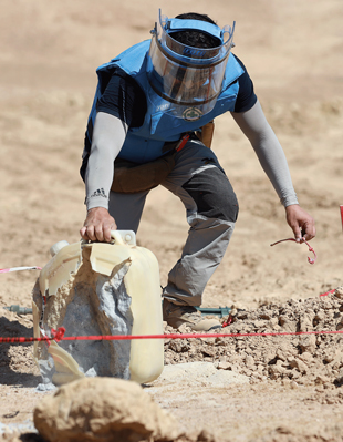 A person in protective gear grabs the handle of a broken plastic container with white powder inside.