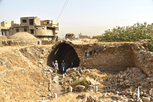 A sandstone arched opening with two people standing in the entrance with debris outside the archway