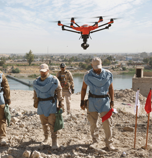 Three people wearing baseball caps and protective vests stand on a beige rocky riverbank looking down at the ground while a red drone hovers above and slightly in front of them.