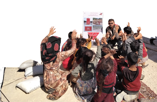 A group of children  with their hands raised in the air sit on rugs layed on a dirt floor looking at an adult sitting next to a large poster with a sign on it that reads Danger Mines.