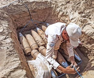 A man in beige clothing squats in a trench containing metal projectiles that are being wired together by the tips with blue wire and silver duct tape.