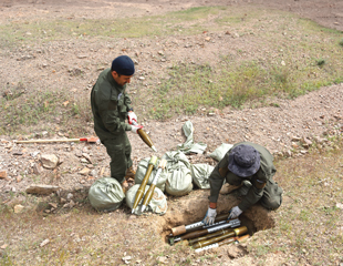 One man kneels beside a hole dug in the dirt ground laying a projectile into a pile of other projectiles in the hold. Another man stands beside him next to sandbags and two projectiles while holding a third.