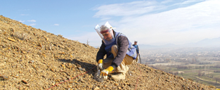 A deminer wearing a blue protective vest and visor kneels on a rocky hillside and digs in the ground with a small trowel.