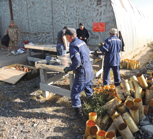 Several people wearing blue jumpsuits work around an outdoor wooded table with boxes of ammunition and projectiles on the ground.