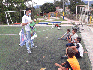 Four children sit on the ground of a soccer field looking a man standing in front of them wearing a mask and holding a poster.