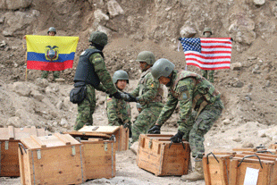Four men in green military uniforms and helmets surrounded by wooden crates place a projectile into one of the crates.
